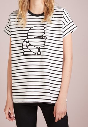 Biała koszulka z czarnymi poziomymi paskami, z czarnym rysunkiem stylizowanej twarzy w profilu na przodzie. Krótkie rękawy.