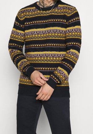 Homme portant un pull en laine noir, jaune et marron à motifs, manches longues, pantalon noir et un collier argenté, mains jointes devant.