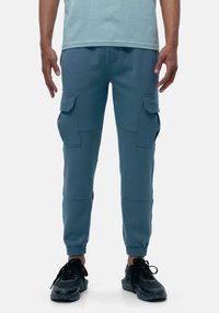 Pantaloni della tuta blu realizzati in un tessuto morbido, con tasche cargo, caviglie affusolate e polsini elasticizzati. Indossati con scarpe sportive nere.