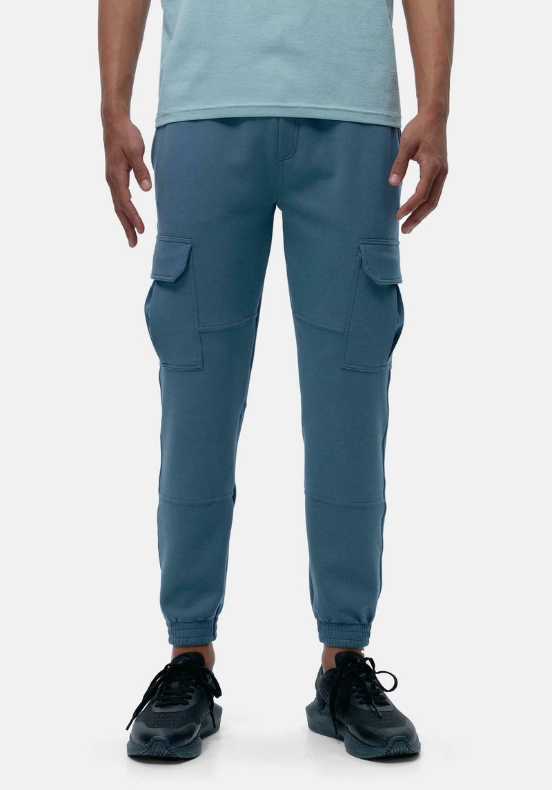 Pantaloni della tuta blu realizzati in un tessuto morbido, con tasche cargo, caviglie affusolate e polsini elasticizzati. Indossati con scarpe sportive nere.