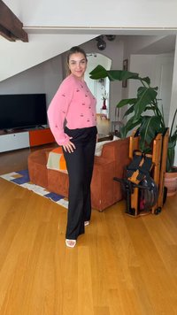 Femme souriante, portant un pull rose à motif cerises et un pantalon noir, debout sur un sol en bois dans un salon avec une grande plante et un appareil d'exercice.