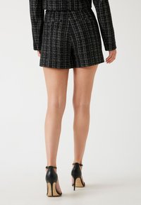 Zwarte geruite shorts met een gestructureerde stof, hoge taille en een getailleerde pasvorm, gecombineerd met zwarte hoge hakken op een lichte achtergrond.
