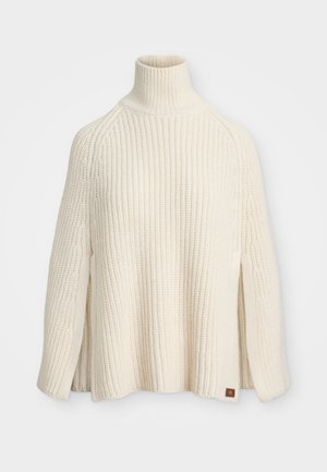 Krems krāsas adīta turtlenecka pončo ar ribotu struktūru, sānu šķēlumiem un mazu logotipa akcentu uz apakšmalas.
