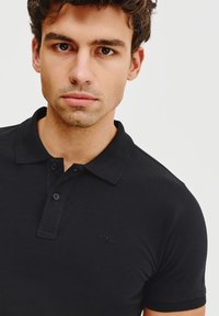 Czarna koszulka polo z klasycznym kołnierzykiem, krótkimi rękawami i subtelnym logo na piersi. Materiał wydaje się gładki i dopasowany.