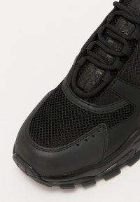 Chaussure de sport noire avec tige en maille et synthétique, détails texturés, col rembourré et semelle en caoutchouc avec motifs de grip.