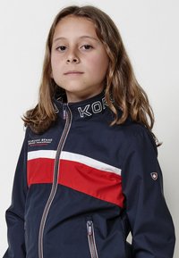 Tengerészi kabát piros és fehér vízszintes csíkokkal, teljes cipzárral, magas gallérral, és logós részletekkel a mellrészen és az ujjon. Sima textúra, szabadtéri stílus.