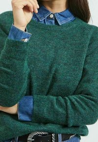 Suéter verde de punto con cuello redondo y mangas largas, superpuesto a una camisa de mezclilla azul con cuello abotonado y un acabado texturizado.