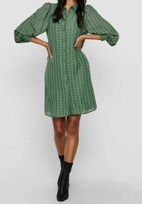 Vihreä raidallinen paitamekko, jossa on napit edessä, lyhyet puffihihat ja rento istuvuus, yhdistettynä mustiin nilkkasaappaisiin. Sileä kankaan rakenne.