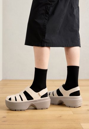 Bežinės platformos sandalai su išpjautomis juostelėmis ir pilku padu. Nešiojami su juodomis kojinėmis, prieš medinį grindų paviršių ir neutralų foną.