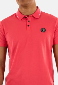 Polo-shirt à manches courtes en corail vif avec col côtelé, patte de boutonnage à trois boutons et un écusson noir sur la poitrine gauche. Tissu lisse.
