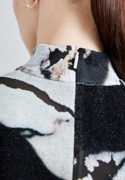 Close-up of a garment collar featuring mixed fabrics; gray with a floral design and black textured section, zip detail at the back.