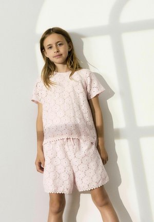Tenue en dentelle rose clair comprenant un haut à manches courtes et un short avec des bords festonnés et des motifs floraux découpés, appuyée contre un mur.