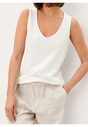 Kobieta w białej koszulce na ramiączkach z dekoltem w serek i beżowych lnianych spodniach, stojąca z jedną ręką w kieszeni na jednolitym tle.