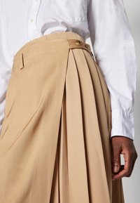 Jupe plissée beige en tissu texturé, dotée d'une taille haute, de poches latérales et d'une fermeture à boutons, associée à une manche de chemise blanche.