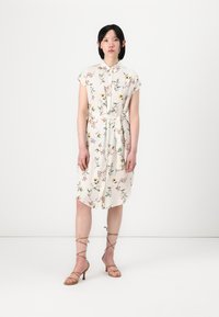 Blumenhemdkleid in Creme mit kurzen Ärmeln und gebundenem Taillenband. Mit grünen Blättern und mehrfarbigen Blumen verziert. Kombiniert mit sandfarbenen Absatzsandalen.
