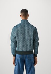 Grön bomberjacka med mönster, hög krage, ribbade muddar och fåll. Har en geometrisk design och kontrasterande sidostrippar.