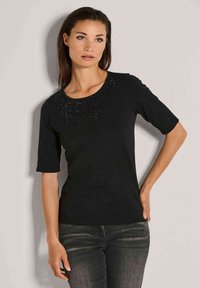 Vrouw met steil bruin haar, gekleed in een zwart t-shirt met korte mouwen en pailletten, en donkergrijze jeans, staand tegen een lichtgrijze muur.