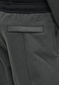 Pantalones de felpa grises con una superficie texturizada, que cuentan con un bolsillo trasero, costuras sutiles y un pequeño logo acentuado cerca de la cinturilla.