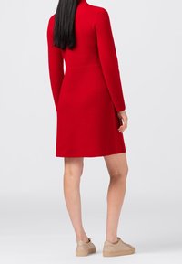 Donna con lunghi capelli neri, vestita con un abito rosso al ginocchio e sneakers beige, in piedi di spalle su uno sfondo neutro.