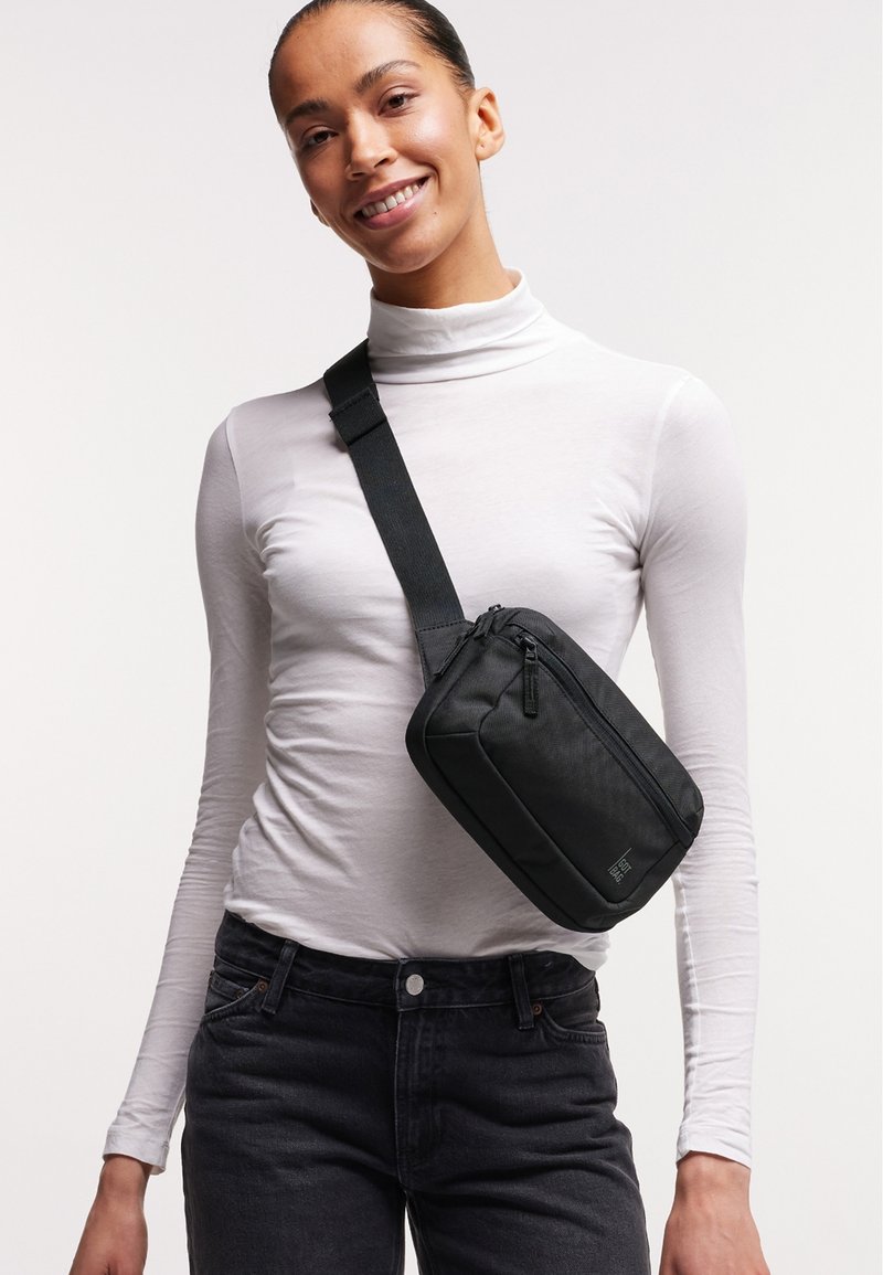 Borsa a tracolla nera in tessuto con chiusura zip, forma rettangolare e tracolla regolabile, indossata su un dolcevita bianco e jeans scuri.