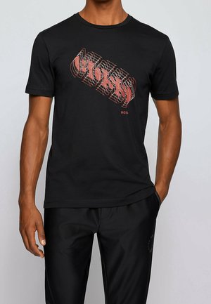 Man draagt een zwart T-shirt met korte mouwen met een roze vervormd "BOSS"-logo op de borst en bijpassende zwarte broek.