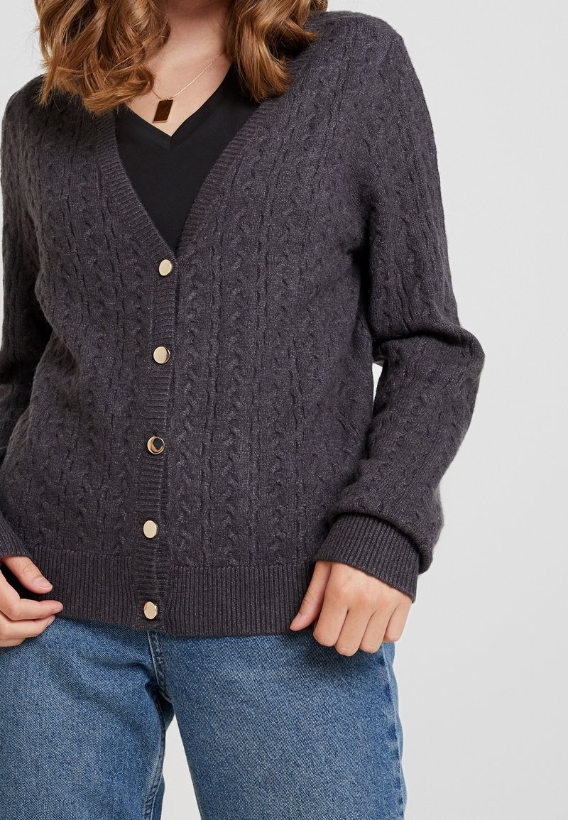 Femme portant un cardigan boutonné gris foncé en maille torsadée par-dessus un top noir et un jean bleu, avec des cheveux bouclés et un collier pendentif en or.