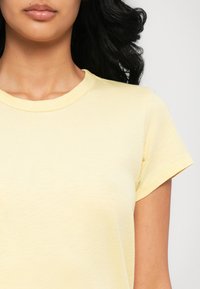 Femme au teint moyen portant un t-shirt jaune clair à manches courtes et col rond, avec des cheveux foncés et ondulés visibles sur le côté droit.