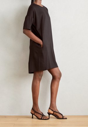 Mujer con un vestido suelto de color marrón oscuro hasta la rodilla con mangas cortas y sandalias marrones de tacón con tiras, de pie contra una pared lisa.