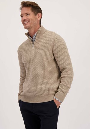 Homme souriant portant un pull quart de zip beige texturé sur une chemise à carreaux, les mains dans les poches, se tenant devant un fond blanc uni.