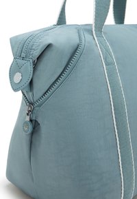 Light blue fabric bag with a zip closure, rounded shape, flat handles, and white accent stripe. Features a leather tag with logo detail.