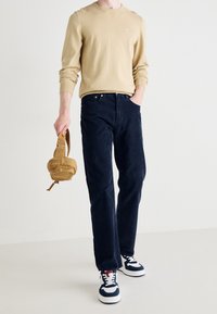 Maglione beige con scollo a girocollo, pantaloni in corduroy blu navy e sneakers blu con accenti bianchi. Tiene una borsa a spalla color ocra.