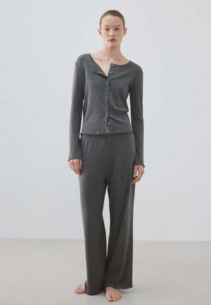 Femme à la peau claire et aux cheveux attachés, debout pieds nus, portant un haut à manches longues gris foncé boutonné et un pantalon large assorti.