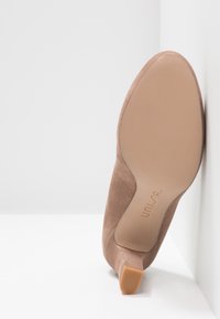 Chaussure à talon haut en daim nude, avec un bout pointu et un talon fin. La semelle est lisse avec un motif texturé. Marquée du logo de la marque.