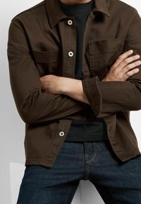 Hnědá kožená bunda s knoflíky a kapsou na prsou, vyrobená z odolné látky. Nositá přes černou košili a doplněná tmavými džíny.