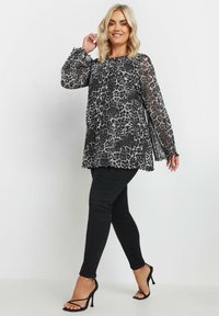 Leopardmönstrad topp i genomskinlig svart och grått tyg med långa, flödande ärmar och volanger vid ärmsluten, kombinerad med svarta slim fit jeans och remsandaler.