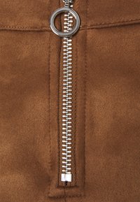 Braunes Kunstsuede mit einem metallischen silbernen Reißverschluss, der einen kreisförmigen Ziehgriff hat, zentral vor einem strukturierten Hintergrund platziert.