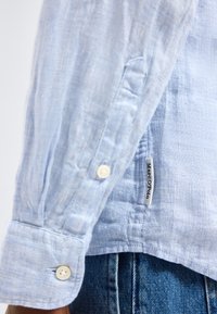 Šviesiai mėlynos ilgomis rankovėmis marškinio rankogalis su baltais mygtukais ir matoma Marc O'Polo prekės ženklo etikete virš mėlynų džinsų.