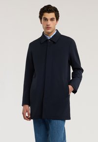 Marineblauer wasserabweisender Mantel mit Kragen, zwei Seitentaschen, vorderer Knopfleiste und geradem Schnitt, getragen über blauem Denim.