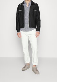 Zwart jack met ritssluiting en witte bies, blauw overhemd met patroon eronder, witte broek en beige sneakers. Eenvoudige, getailleerde pasvorm met strakke lijnen.