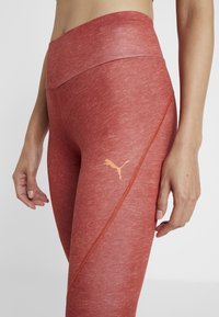Leggings athlétiques rouges avec une texture chinée, une taille haute et des détails de couture latérale. Comprend un logo Puma orange sur la cuisse.