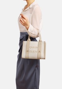 Borsa beige con lettering dorato "QUEEN HELENA", forma rettangolare, tessuto texturizzato, doppio manico superiore, presentata su uno sfondo chiaro.