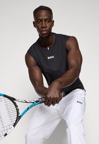 T-shirt sans manches noir avec le logo "BOSS", associé à un pantalon blanc et tenant un racket de tennis avec un design bleu et noir.