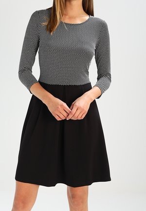 Donna che indossa un abito nero con una parte superiore a maniche lunghe aderente e decorata e una gonna nera semplice, con le mani incrociate in vita.