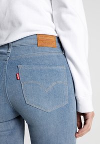 Light blue Levi’s jeans with a leather brand patch and red tag, worn with a white long-sleeve shirt, showing back pocket detail.