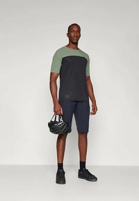 Kortermet T-skjorte med grønn overdel og svart underdel, kombinert med marineblå shorts. Modellen holder en svart sykkelhjelm og har på seg svarte joggesko.