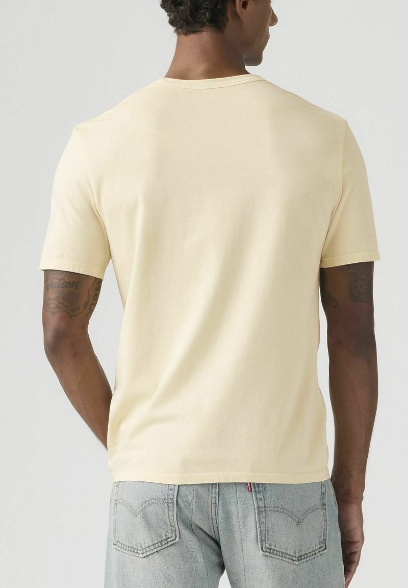 Uomo che indossa una t-shirt gialla chiara a maniche corte e jeans azzurri, visto di spalle su uno sfondo neutro.