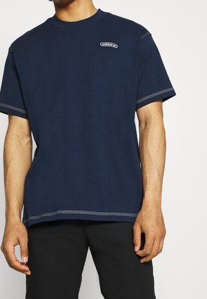 Mężczyzna ubrany w granatową koszulkę Adidas z białymi przeszyciami i czarne spodnie, pokazany od klatki piersiowej do połowy uda na jednolitym tle.