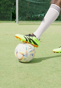 Botín de fútbol verde brillante con acentos negros, colocado sobre un balón de fútbol multicolor en un campo de césped verde. Medias blancas visibles.