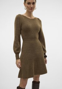 Abito a maglia marrone con corpetto aderente, maniche lunghe a sbuffo e gonna svasata, realizzato in un morbido tessuto texturizzato. Accentuato con orecchini a cerchio dorati.