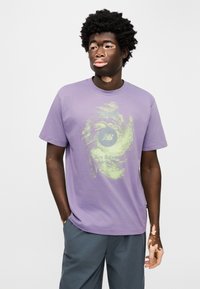 Μωβ βαμβακερό T-shirt με πράσινο γραφικό τύπωμα και κείμενο από τη New Balance. Κοντά μανίκια, στρογγυλή λαιμόκοψη, άνετη γραμμή.